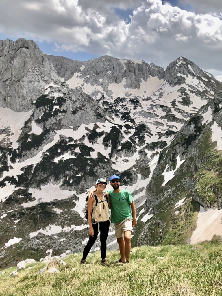  Caroline, in a yellow top and black pants and Jonathan, in a green shirt and blue hat, stand on a grassy hill against snow covered mountain backdrop. 