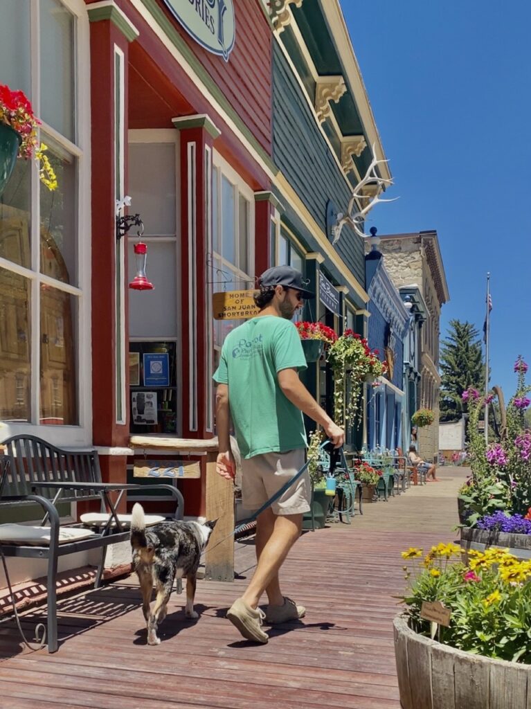 Jonathan, in a green shirt and tan shorts, walks on a wooden boardwalk next to flower planters and historical & colorful buildings. 