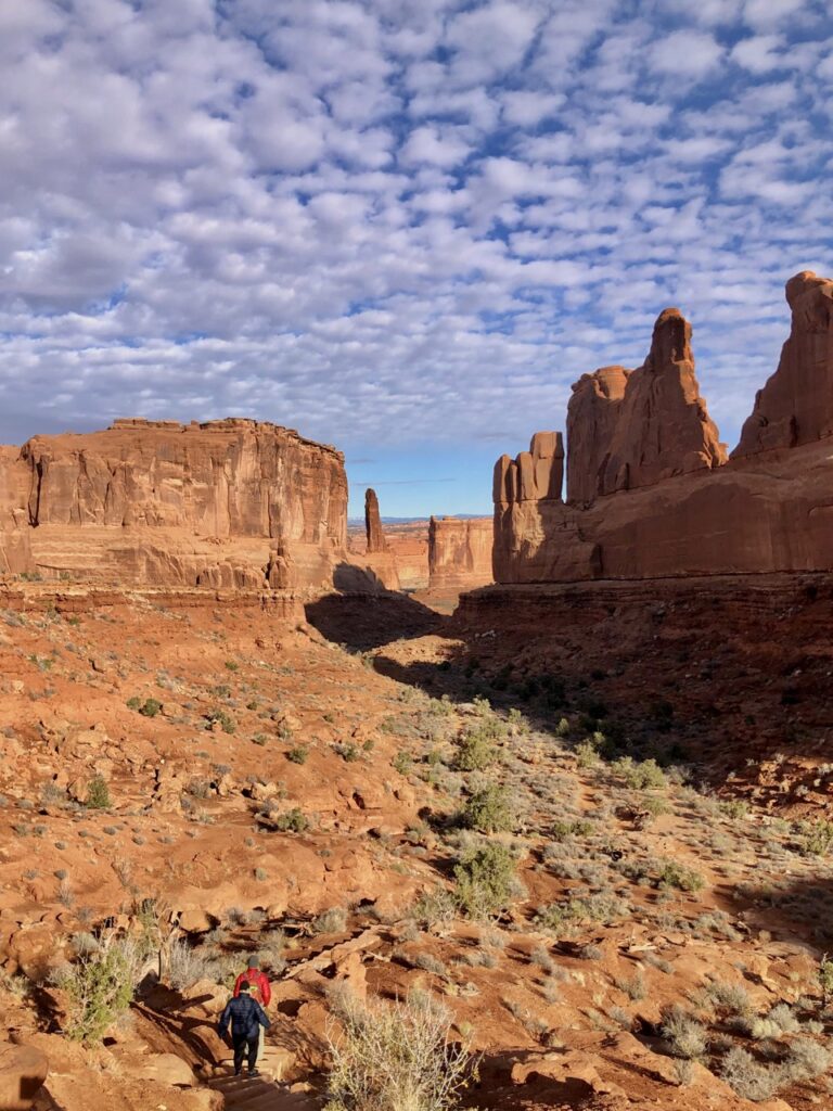 2 small hikers at the bottom of the screen follow a trail in a canyon of two towering red rocks. blue sky with dotted white clouds. 