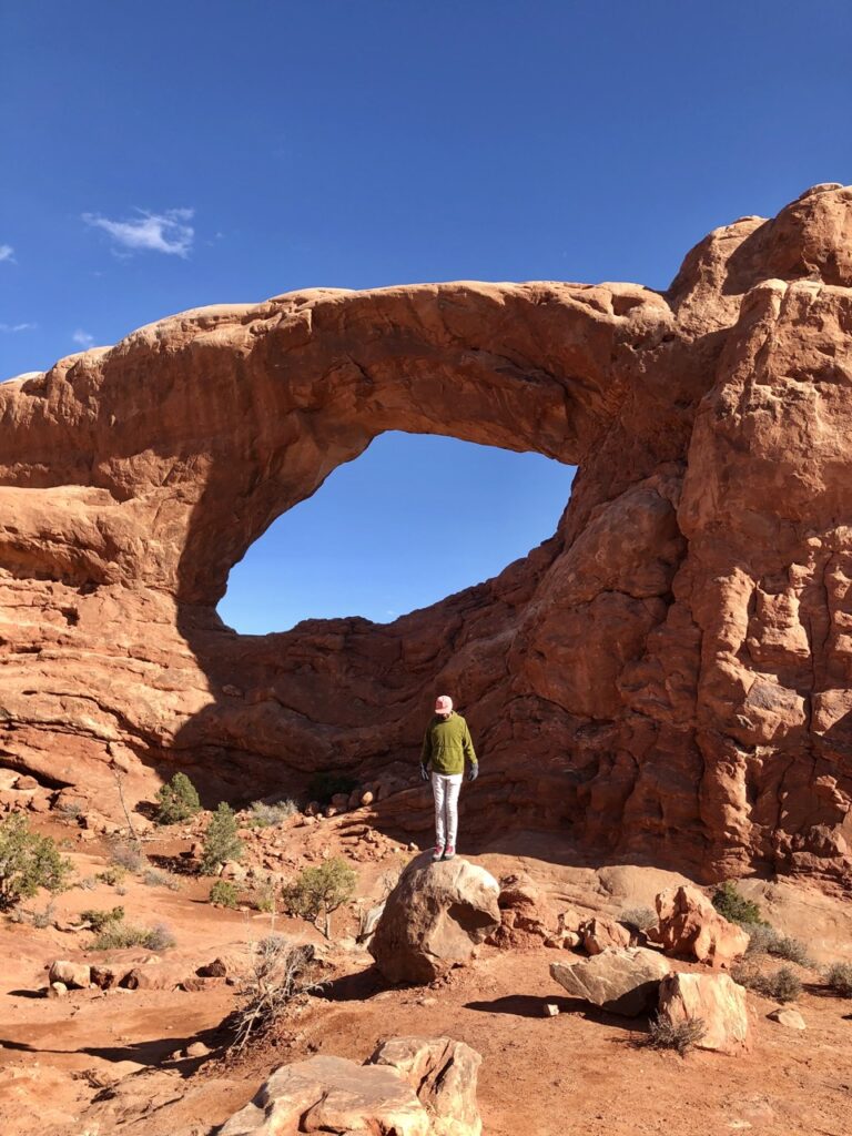 Jonathan, in a green sweater, stands on a red rock in Arches National Park with a "rock window" and bright blue sky behind him. 