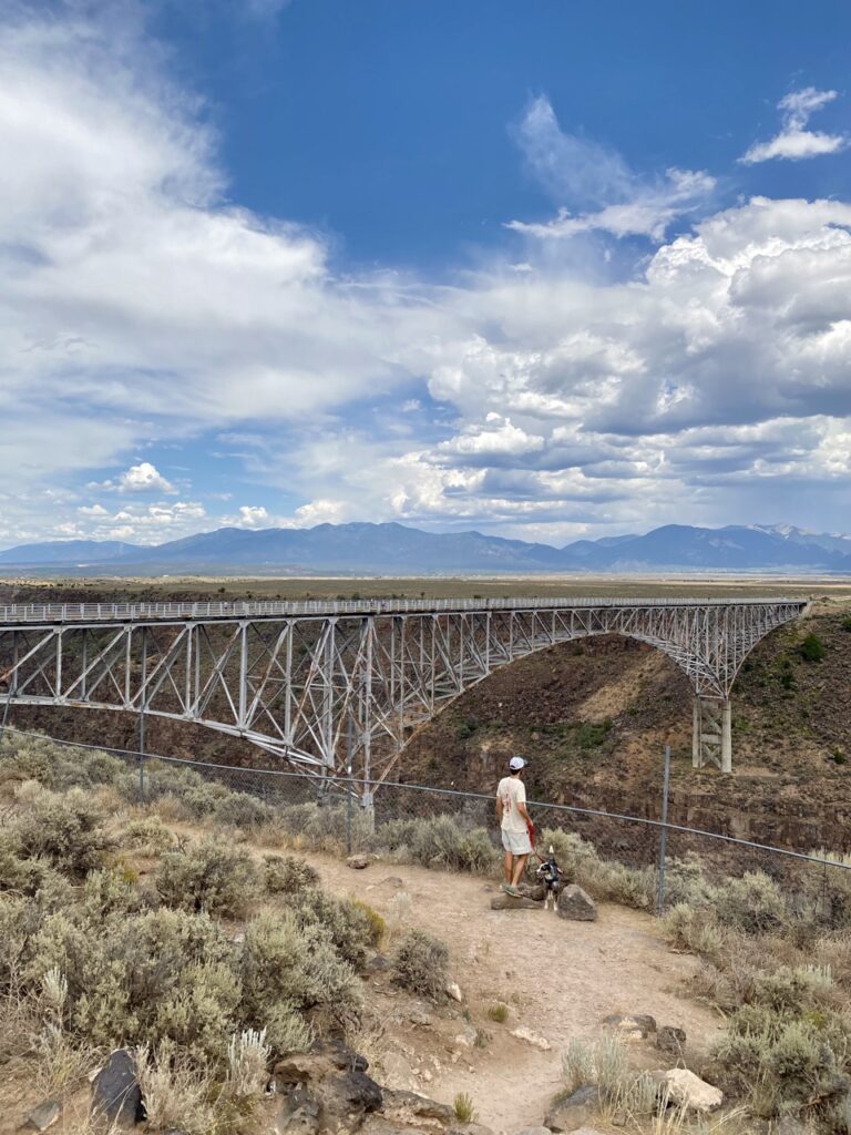 Jonathan, in a white shirt and tan pants, stands at the edge of a gorge looking at a metal bridge. 