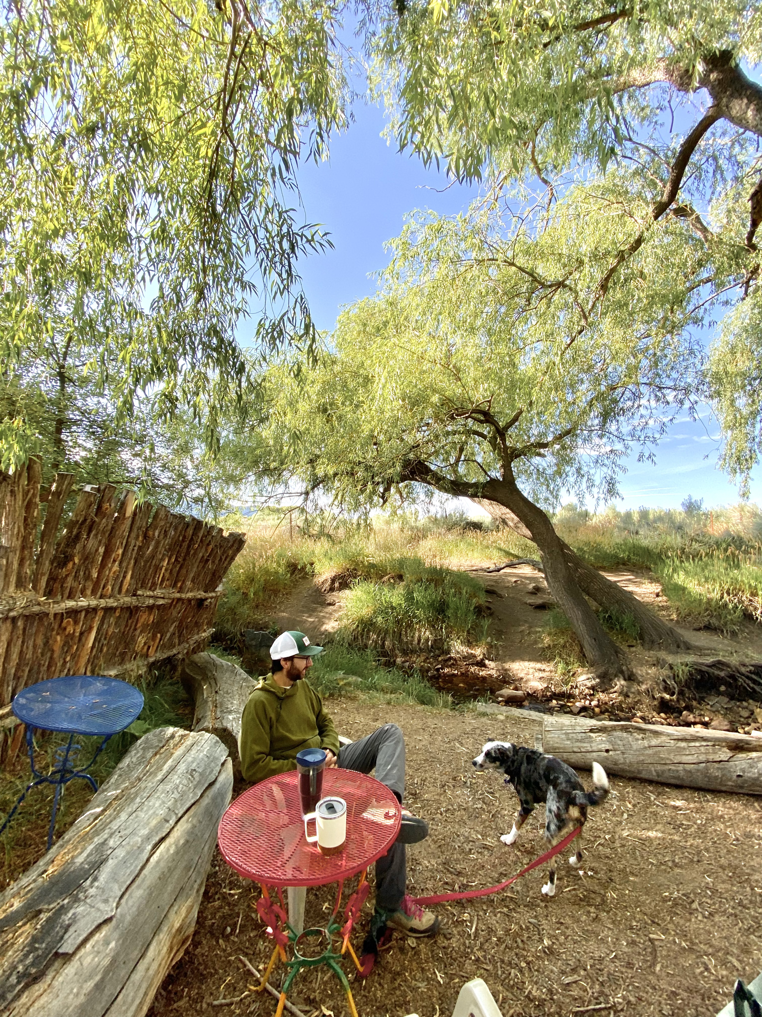 Jonathan, in a green shirt, sits at a red table with coffee cups on it. Behind him are several trees and a small stream. top 7 things to do in taos, new mexico. 