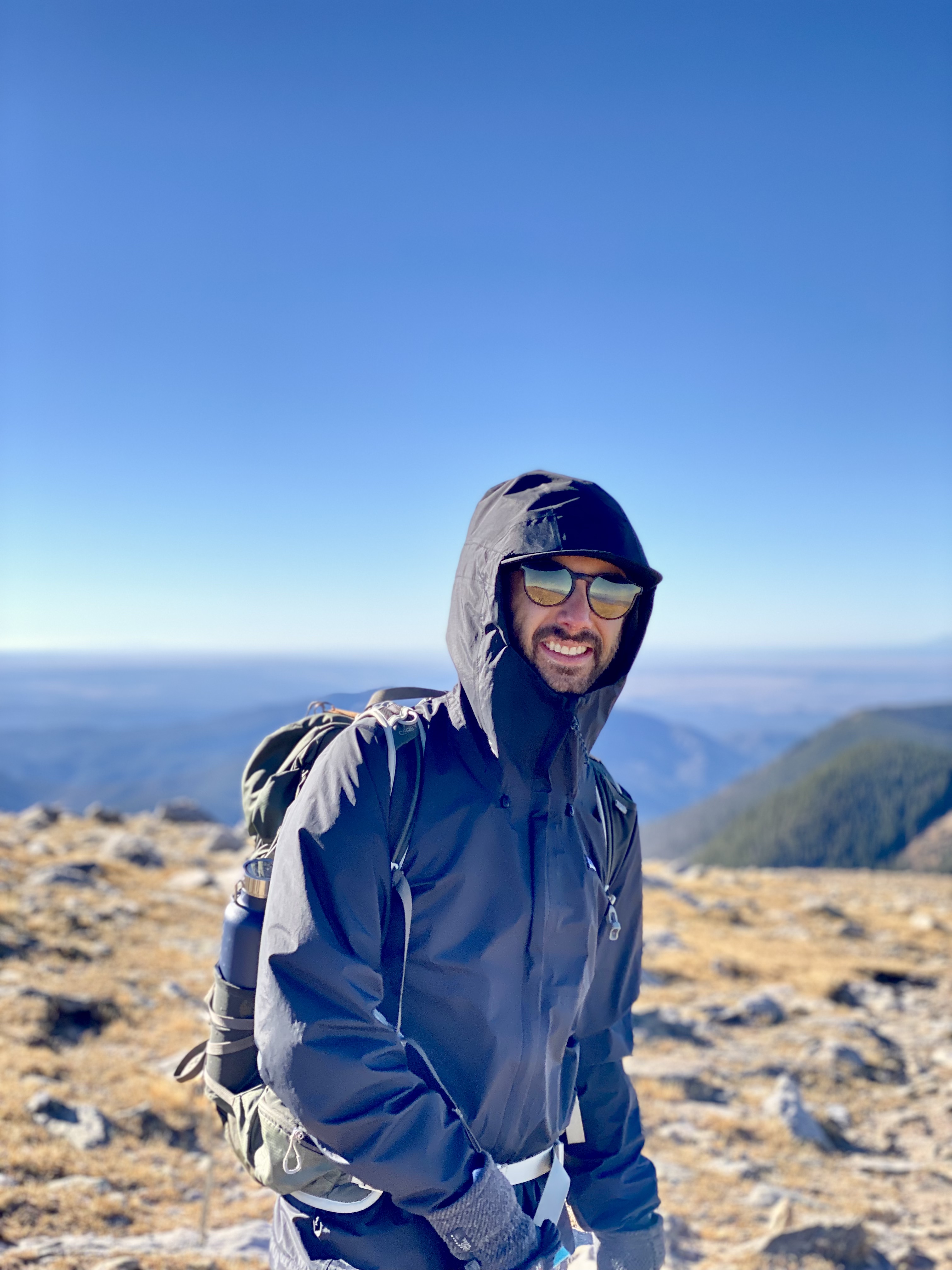 Jonathan, in a black jacket, sunglasses and green backpack, stands on top of a mountain. 