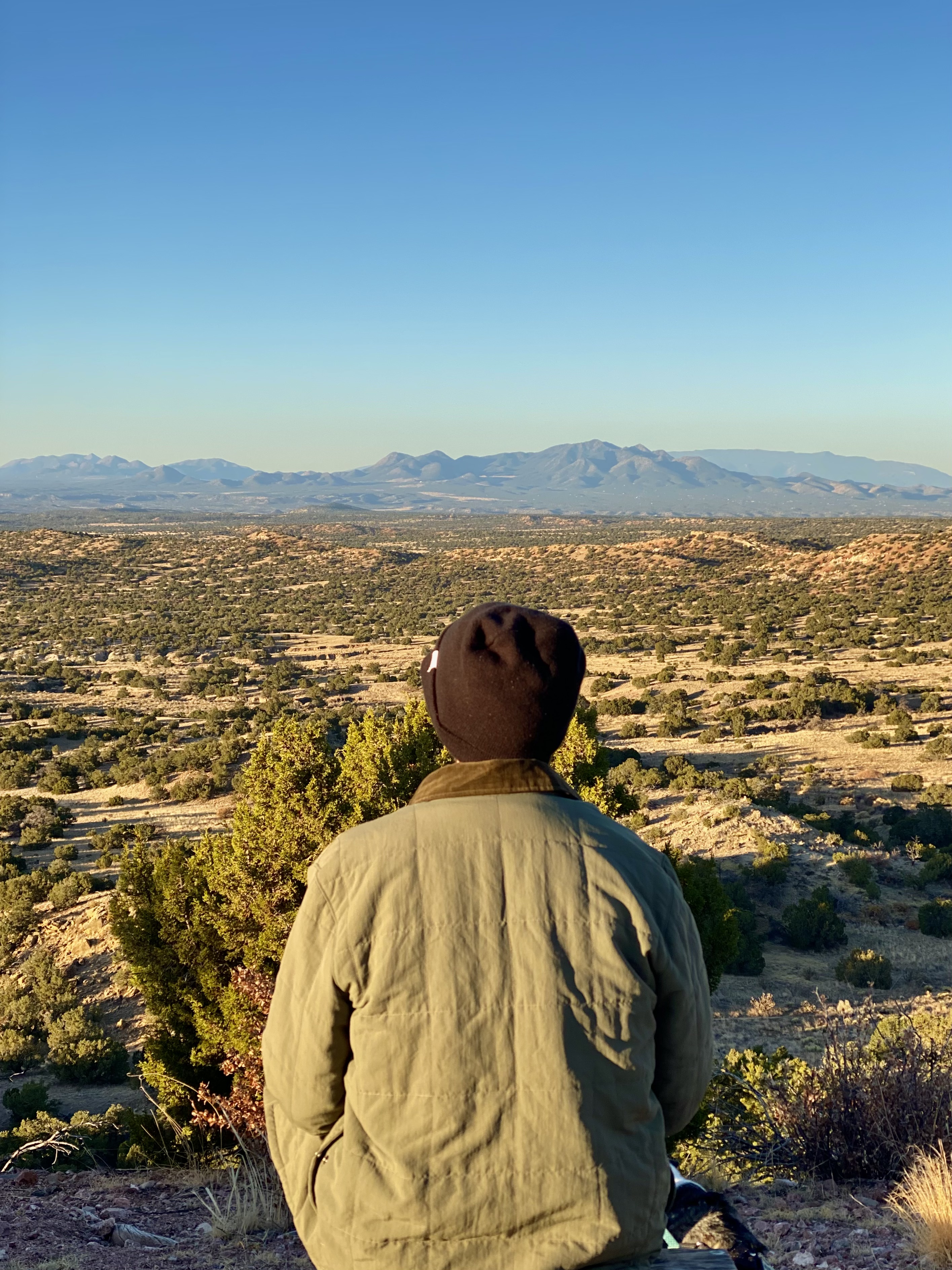 Jonathan, in a green jacket and black beanie, sits on a bench overlooking green juniper trees and distant mountains. 