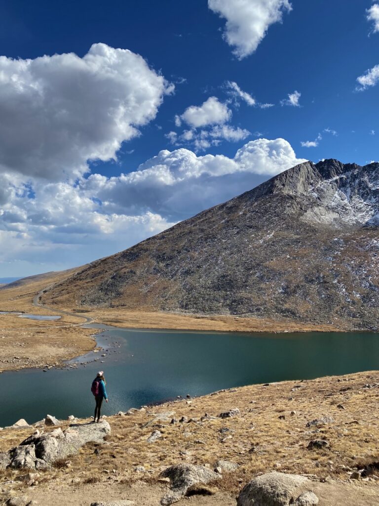 Caroline, standing on a rock with a turquoise jacket, looks at a blue lake and mountain background. 