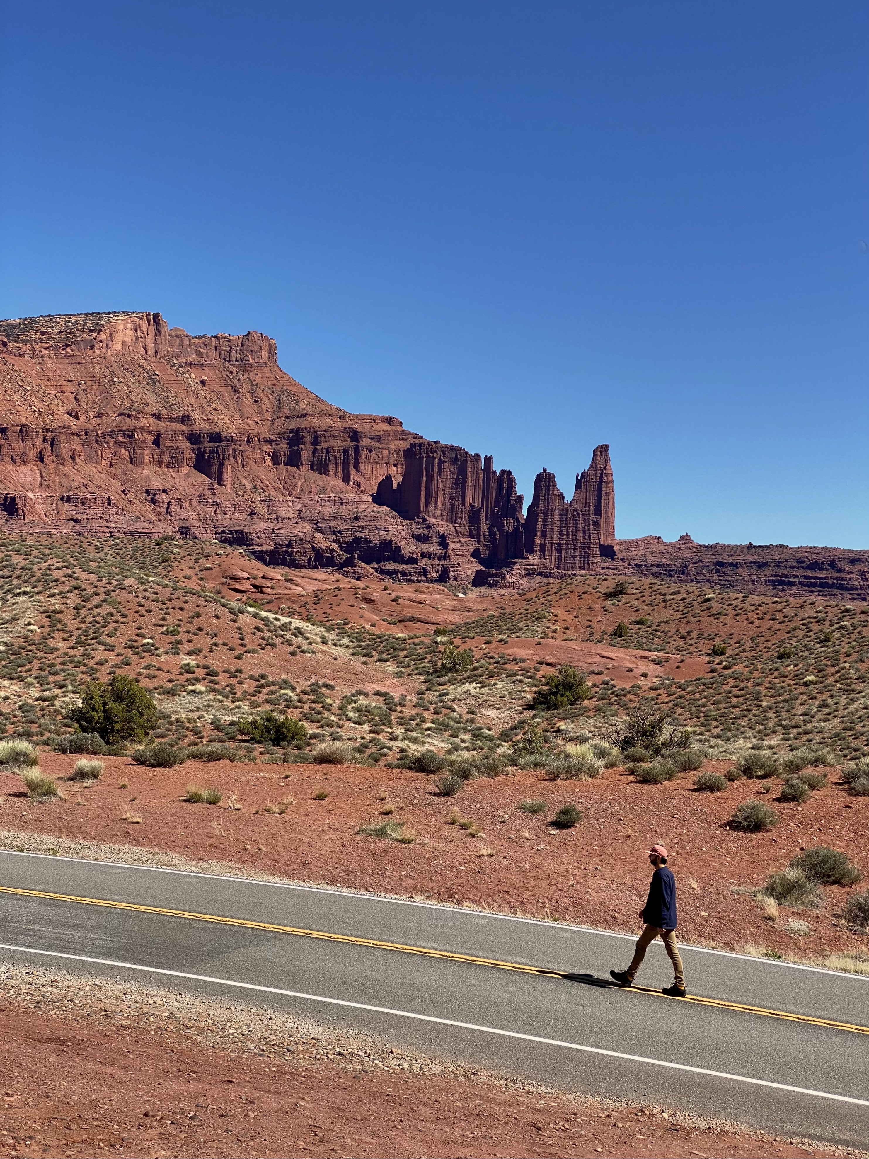 Jonathan in a blue shirt and tan pants walks along the highway with red jagged rock formations in the background and small green plant life. 
