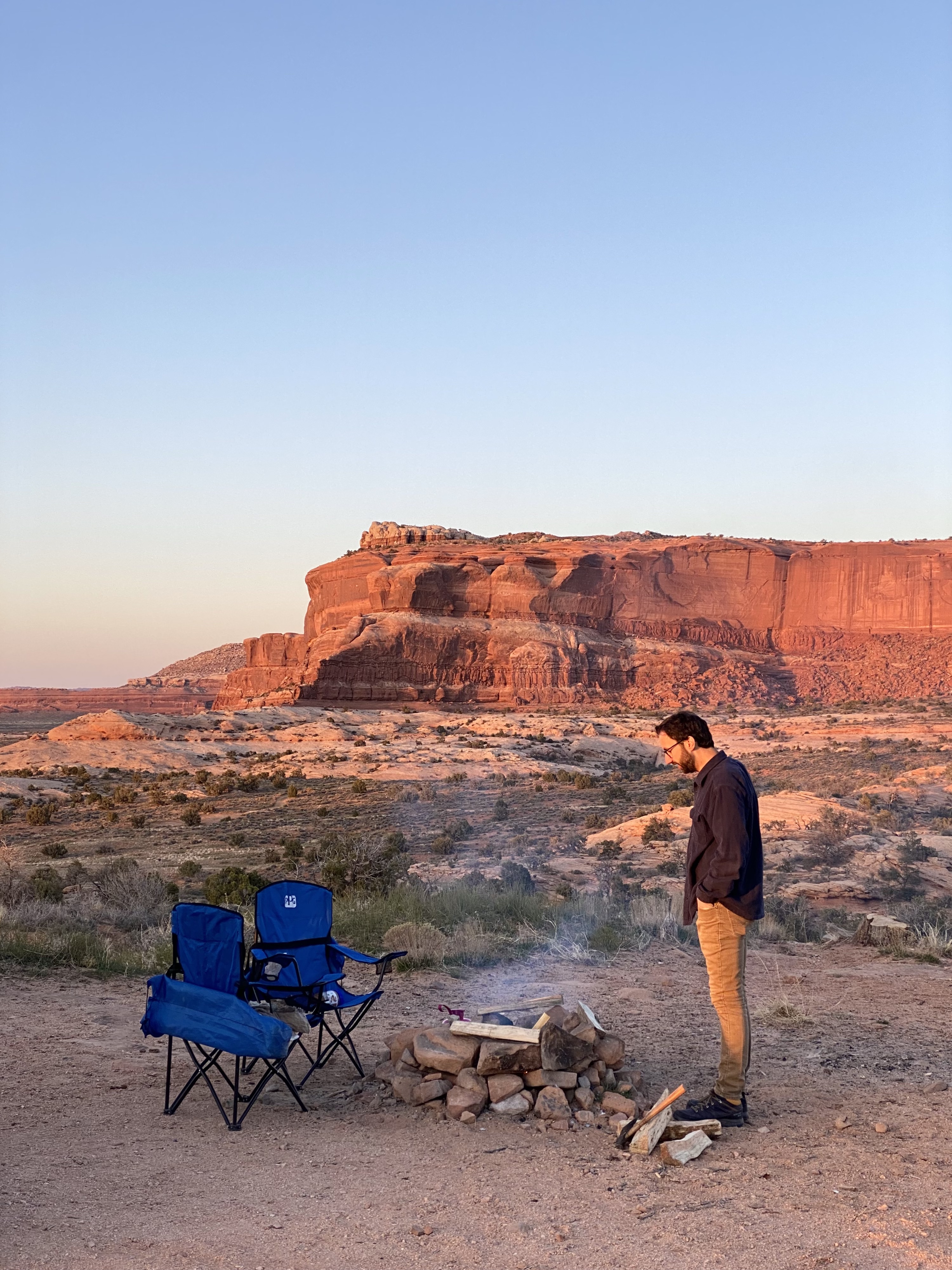 how to spend a weekend in moab, utah. jonathan with a navy shirt and tan pants stands at a fire with 2 blue chairs and sunset red rock in background. 