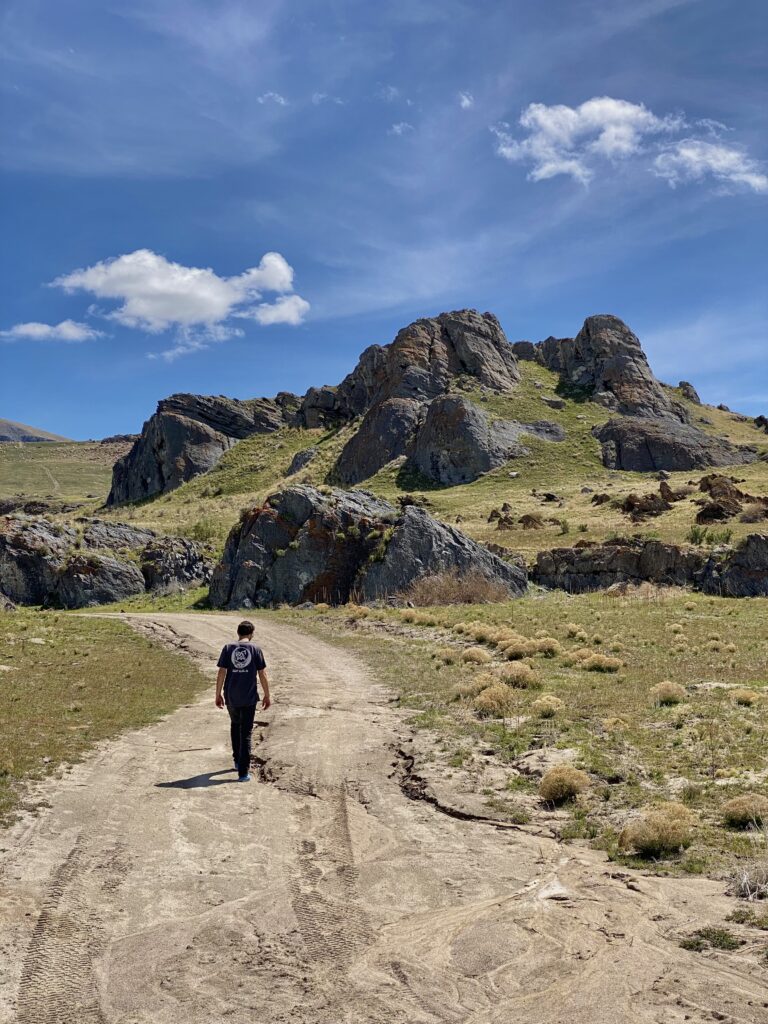 Jonathan walking in a gray shirt and gray pants up to green mountains and gray rocks with blue sky background. 