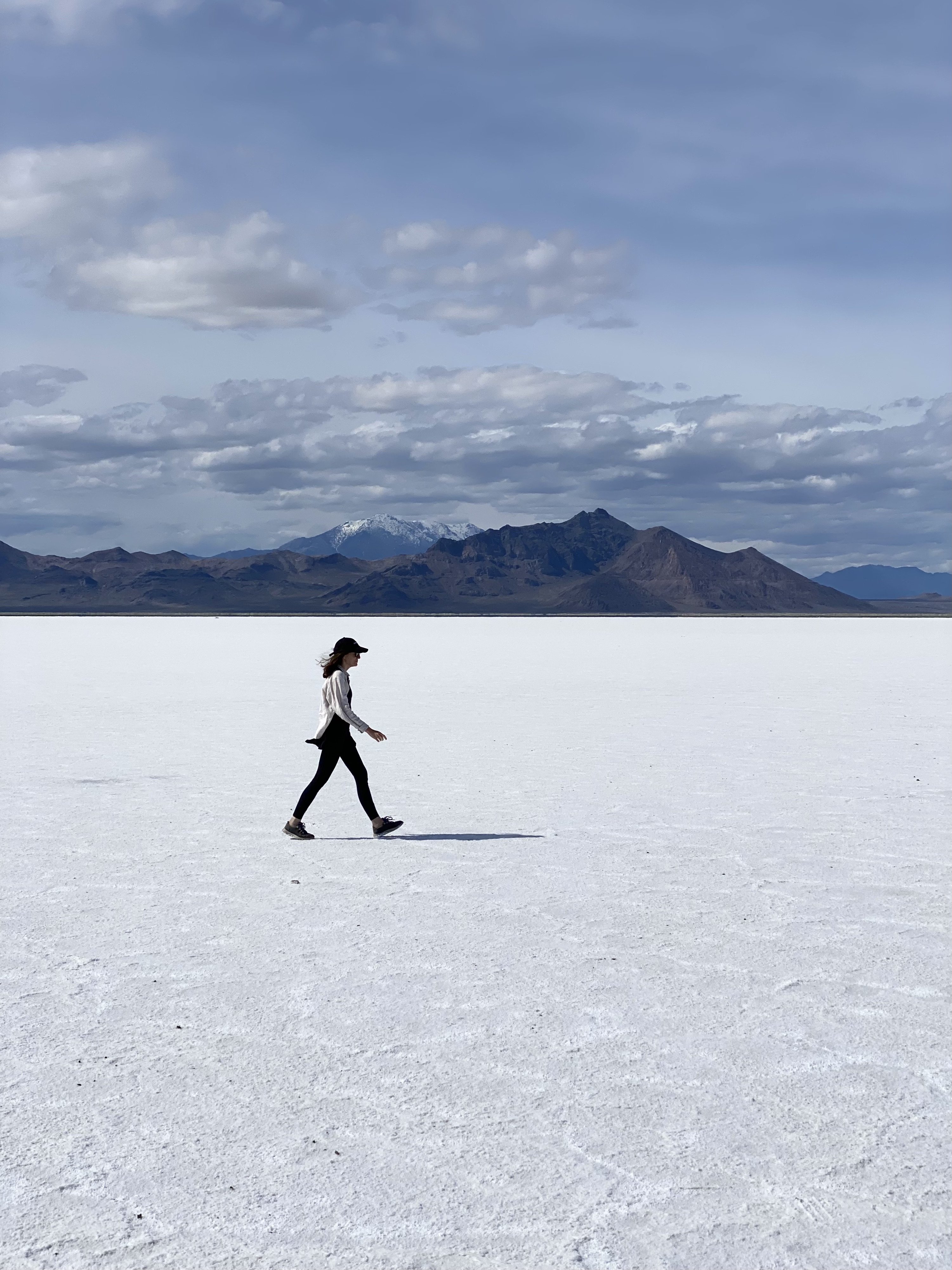 Caroline, in a white shirt and black pants, walks across the bright white salt flats landscape with distant brown mountains in the background. 8 amazing places to visit on a utah road trip.  