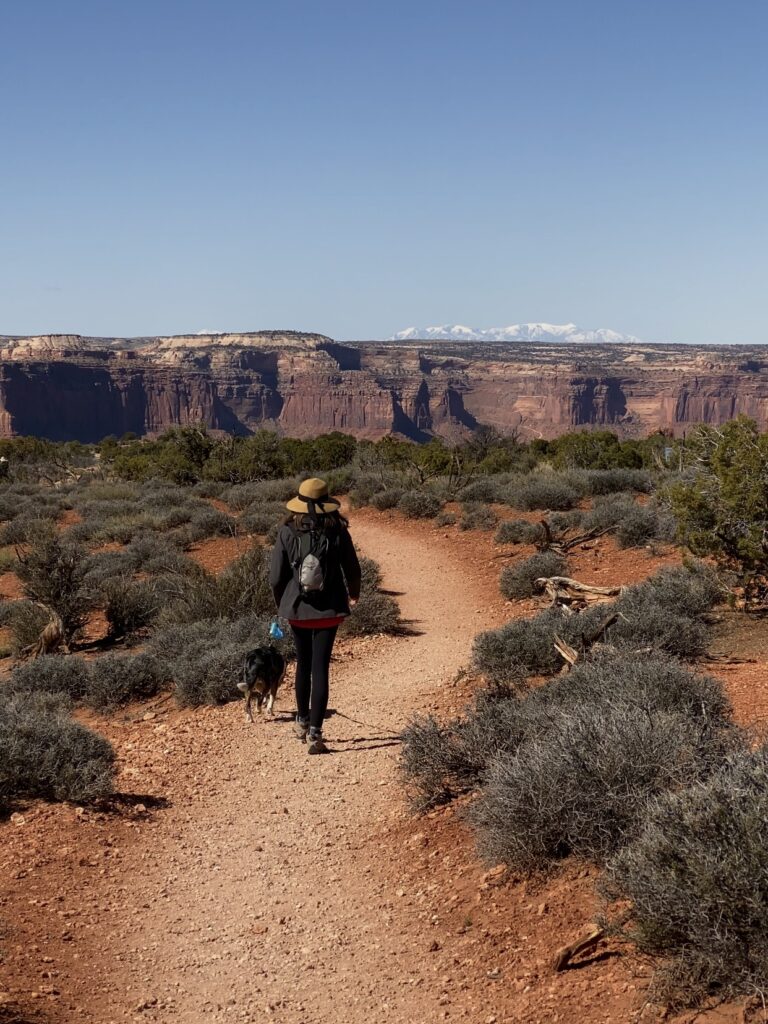 Caroline, in a black jacket and black hiking pants walks across a trail with scrub bush, red dirt and distant canyon. how to spend a weekend in moab, utah