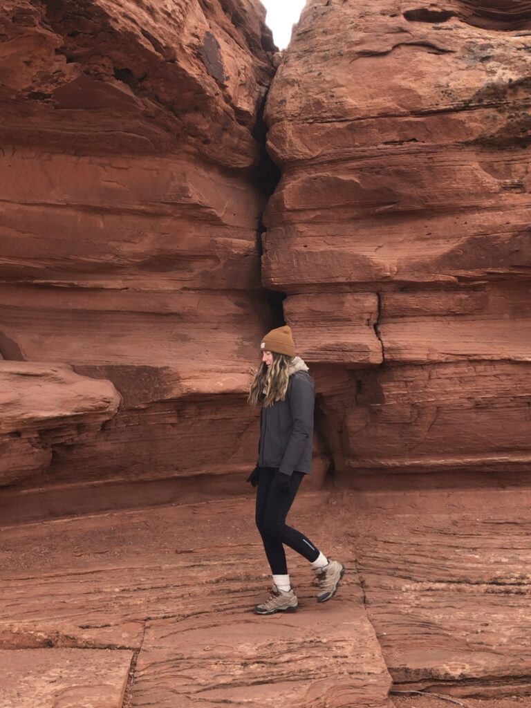 Caroline, in a black jacket and black pants and hiking boots, walks across red rock wall. 