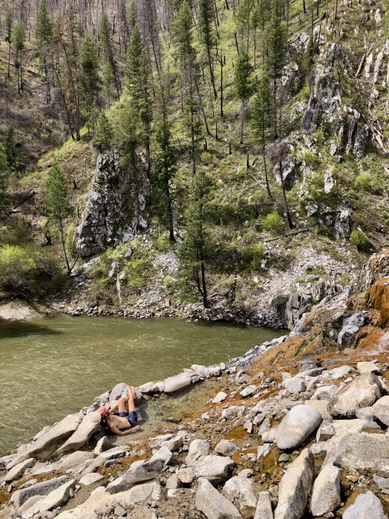 Jonathan, dressed in a bathing suit, sits in a small pool with a green river. mountain, and pine trees in the background. 