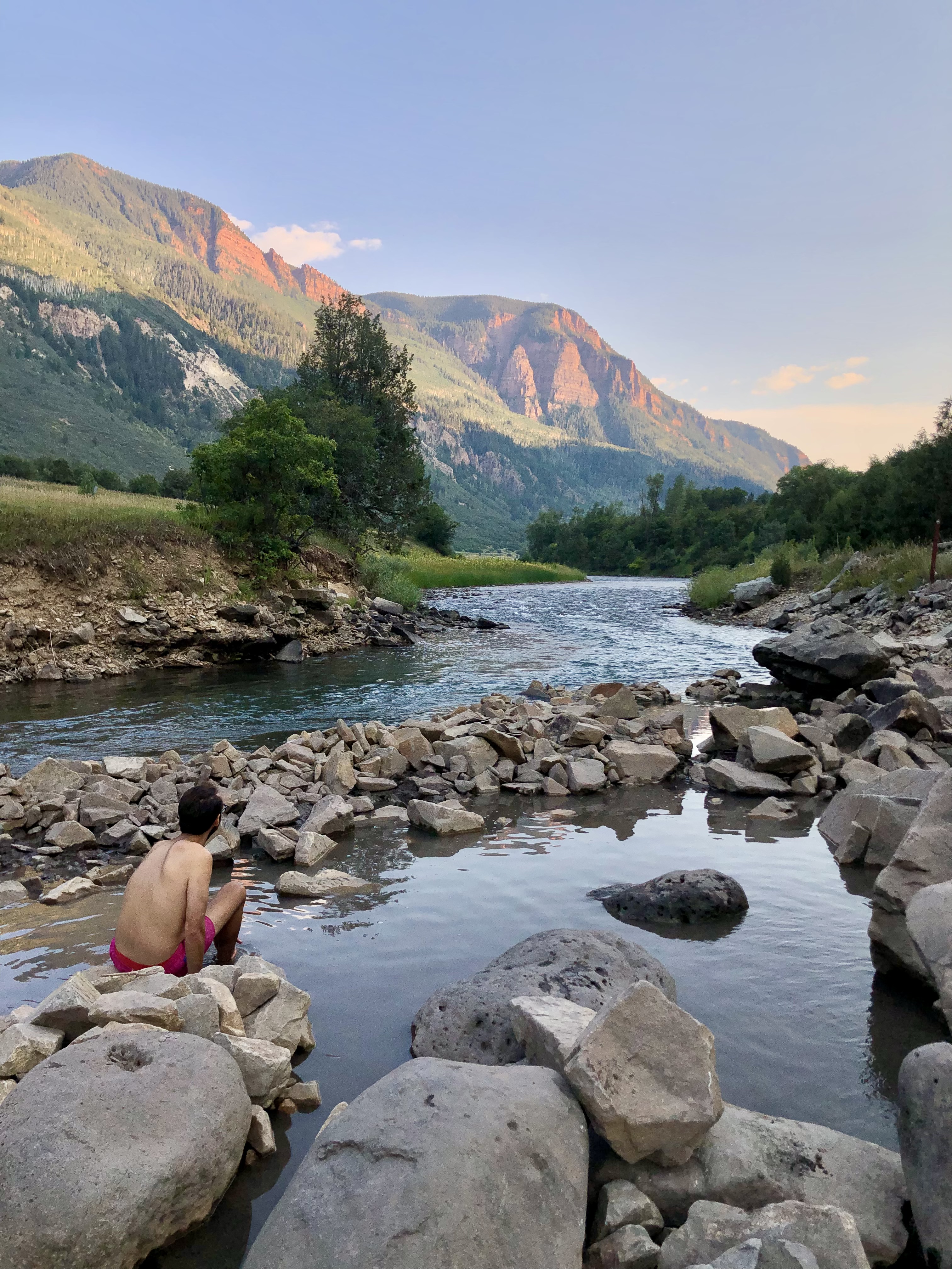 Jonathan, in a pink bathing suit, looks at sunset in the mountains. He sits in a hot spring along a blue river. 