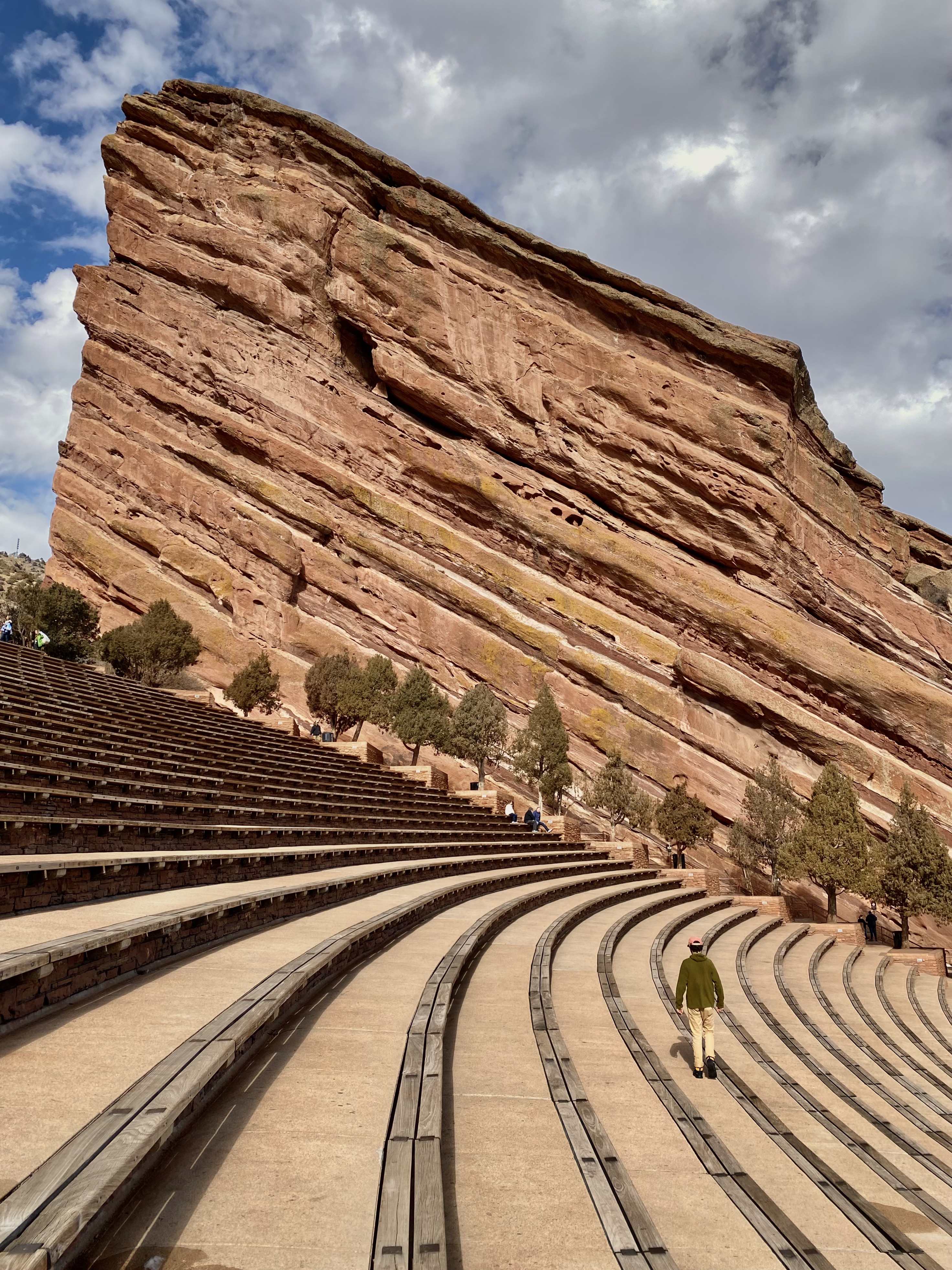 Towering red rock face with amphitheater seating and jonathan, in green sweater and tan pants, walking across them. 10 beautiful spots to visit in colorado (no hiking required)