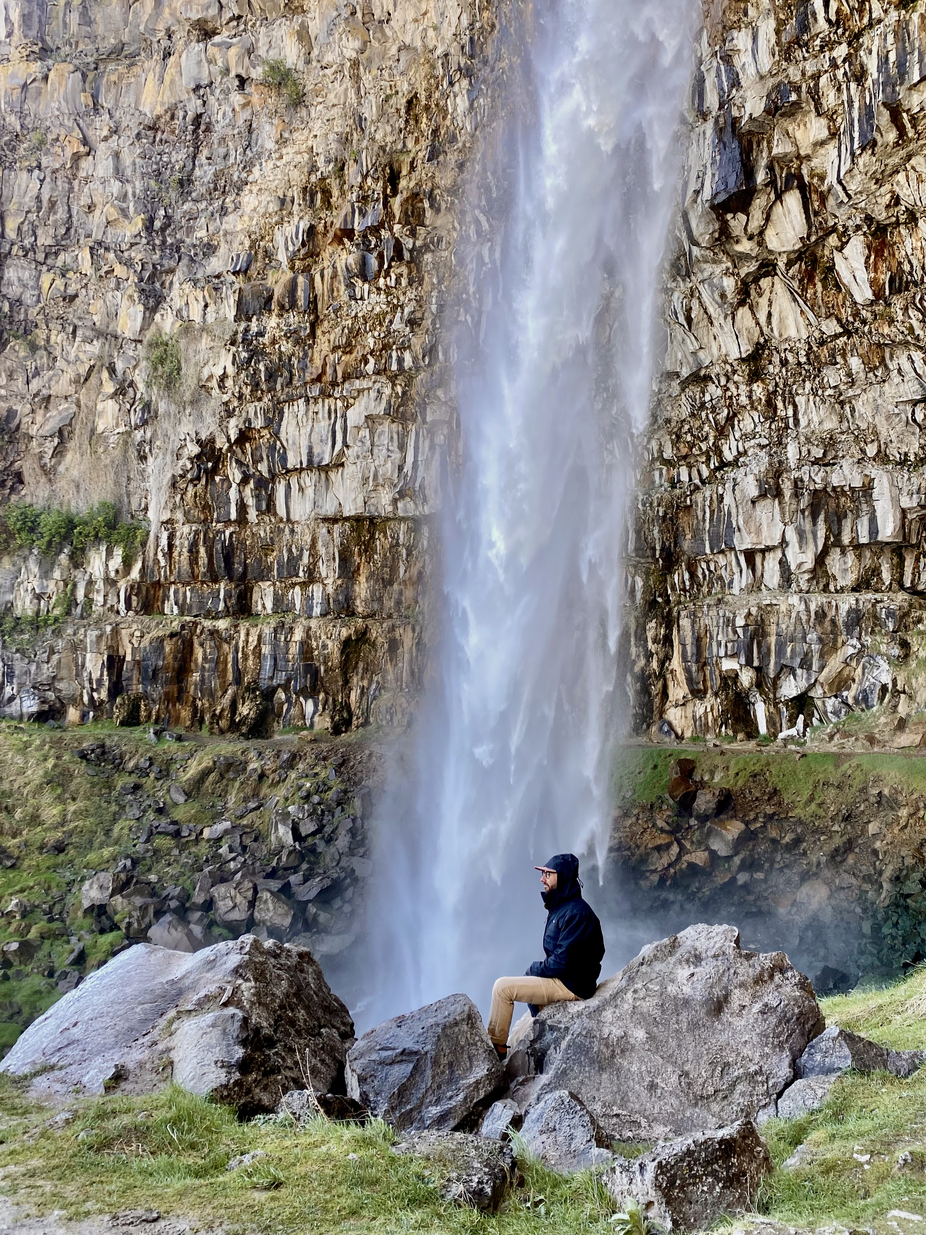 jonathan, in a black rain jacket, sits on a rock in front of a tall waterfall. unique places to visit on a southern idaho road trip. 