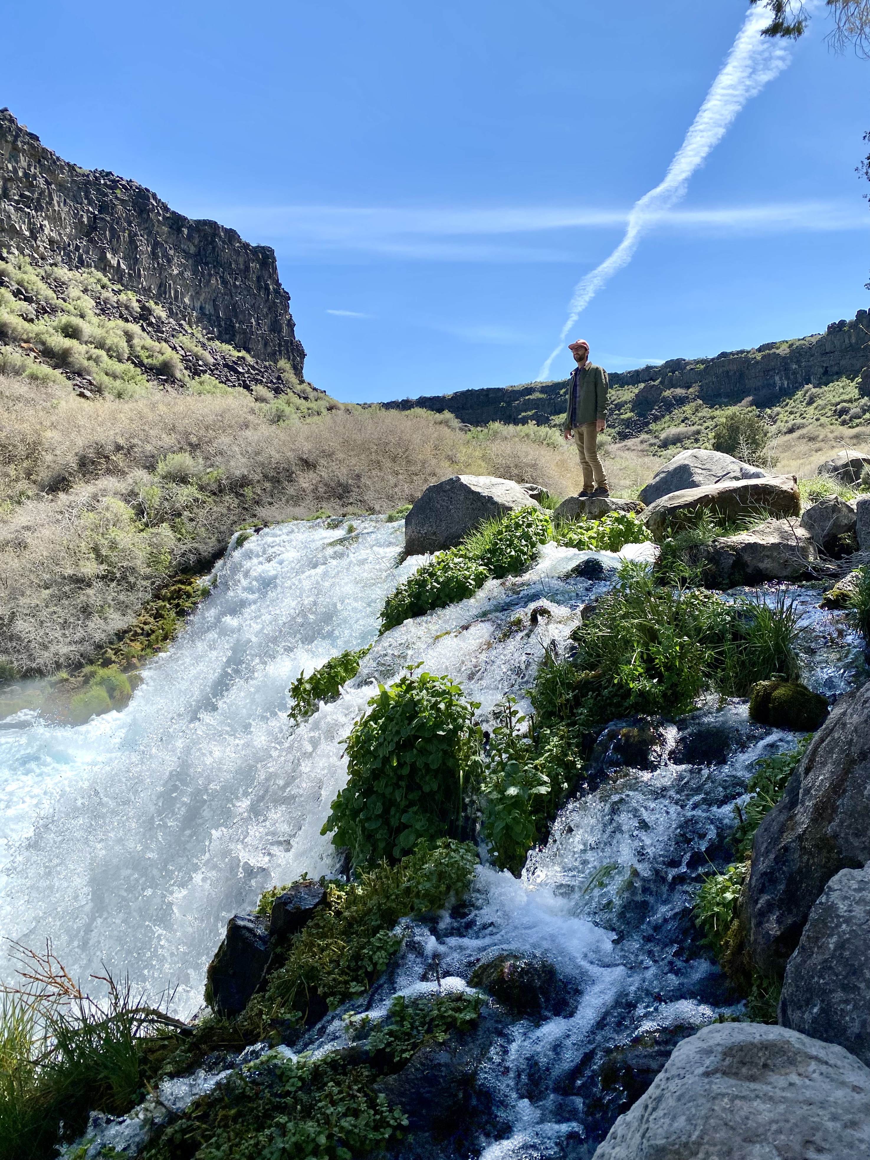 Jonathan, in a green jacket and pink hat, stands on a rock above a flowing waterfall. green moss covered rocks surround the fall and grass.unique places to visit on a southern Idaho road trip.