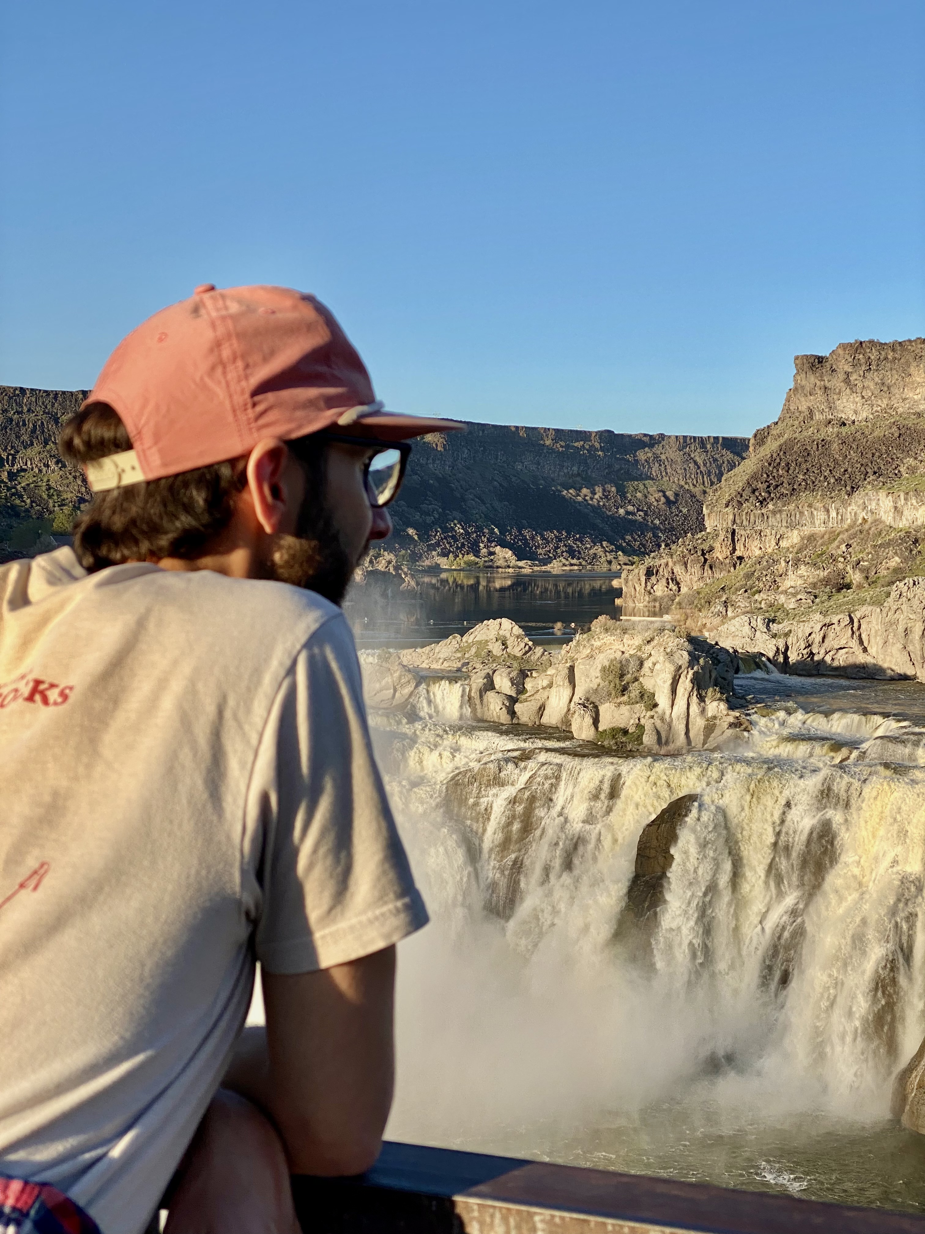 Jonathan, in a beige shirt and pink hat and glasses, looks at a waterfall. amazing places to visit on a southern idaho road trip. 