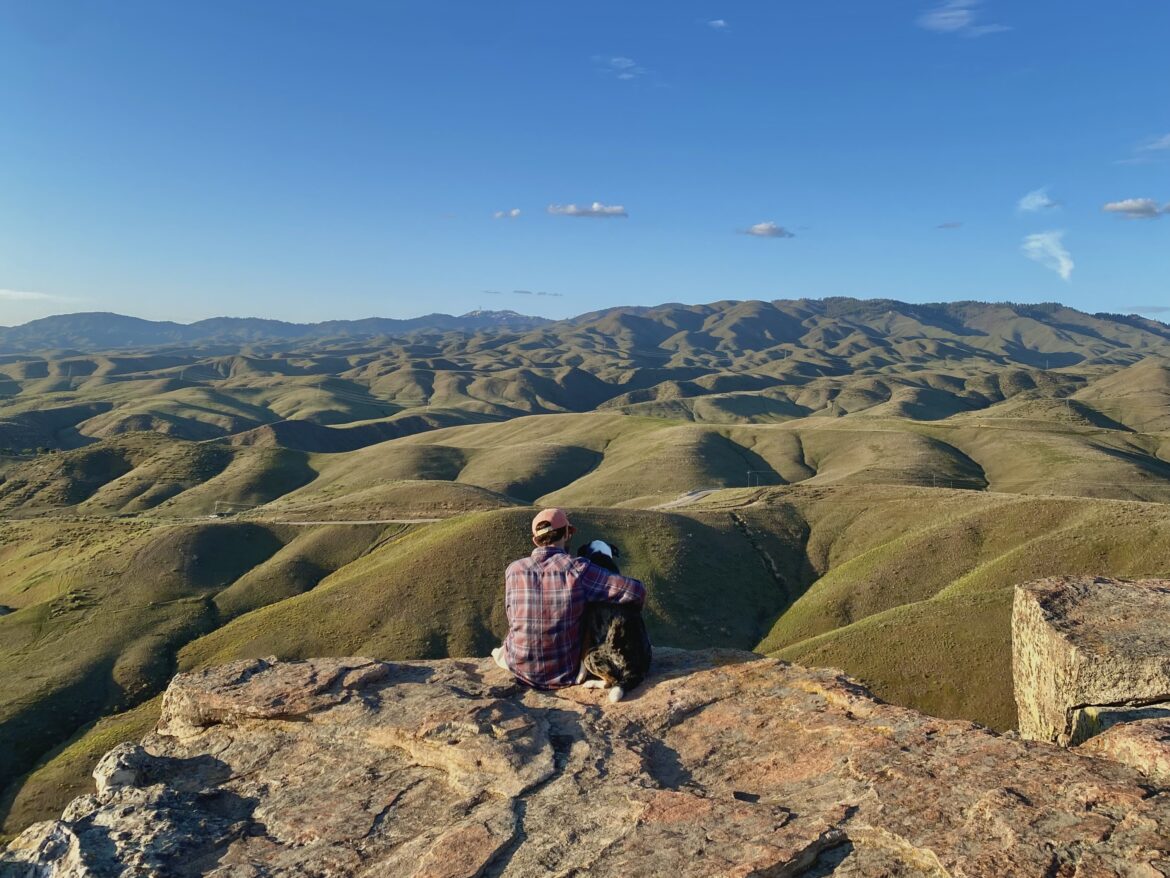 Jonathan sits on the edge of a rock with a pink hat and plaid shirt on. Green grass covered hills form the backdrop behind him