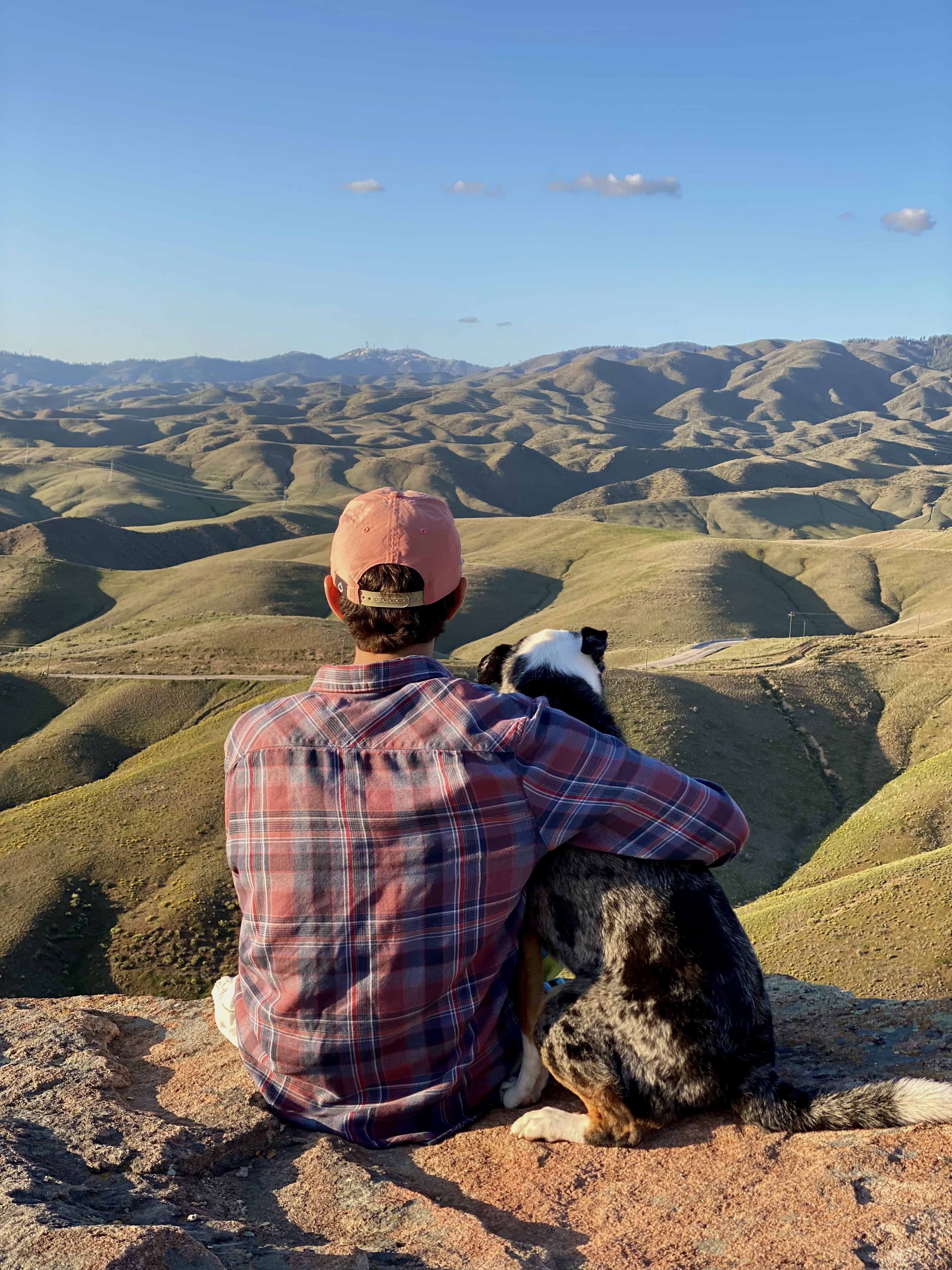 Jonathan, in a pink hat and purple shirt, sits on a rock next to Queso. Queso is a dog with spotted black and white fur. 