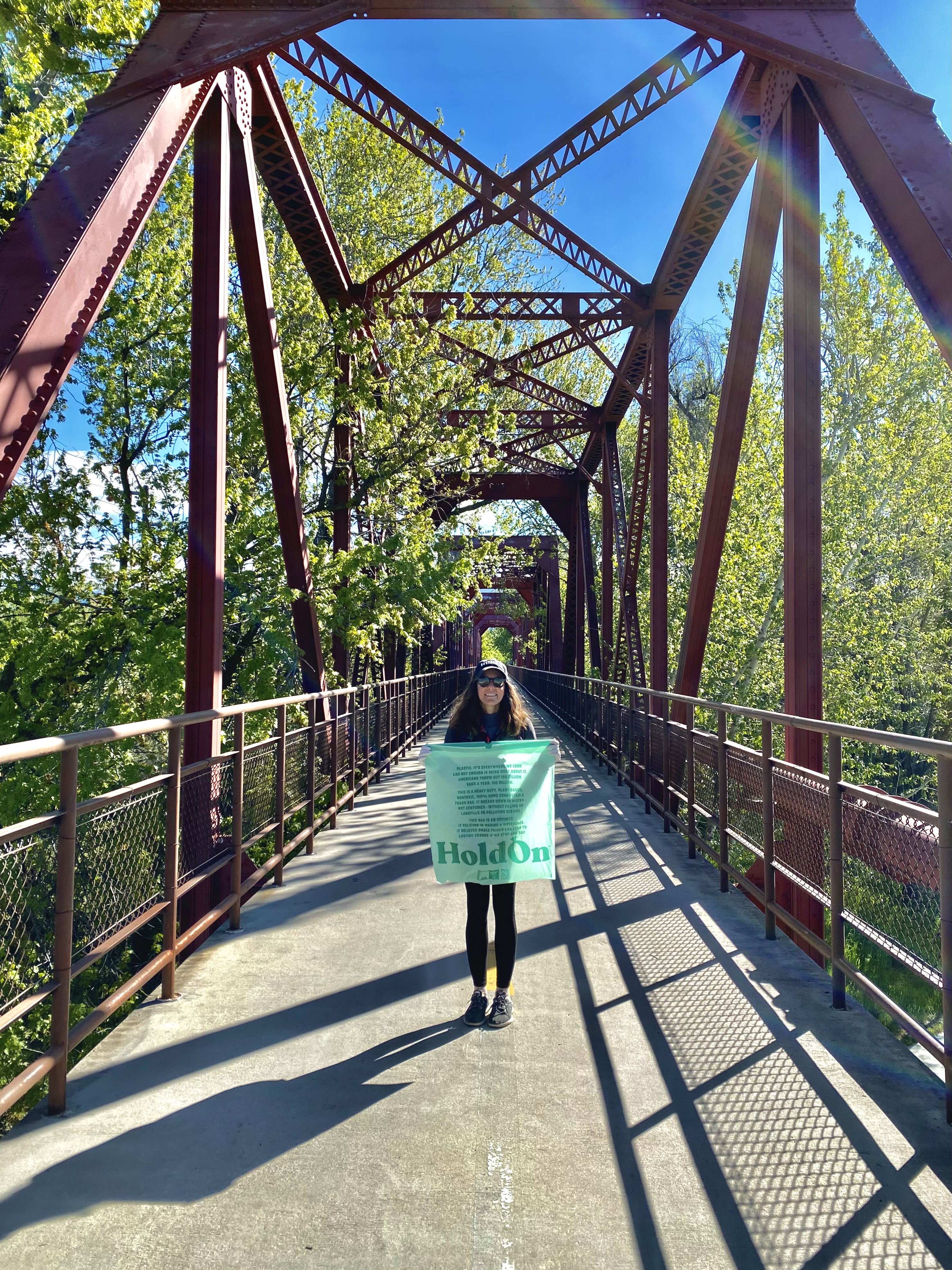 Caroline stands in a black sweater and black leggings on a metal bridge. green trees surround the bridge. outdoor activities around boise