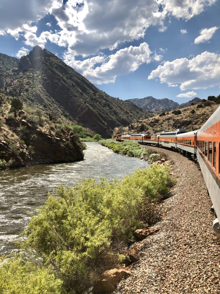 Orange and gray train rides alongside a river, green plants, and mountains. 10 beautiful spots to visit in colorado (no hiking required)