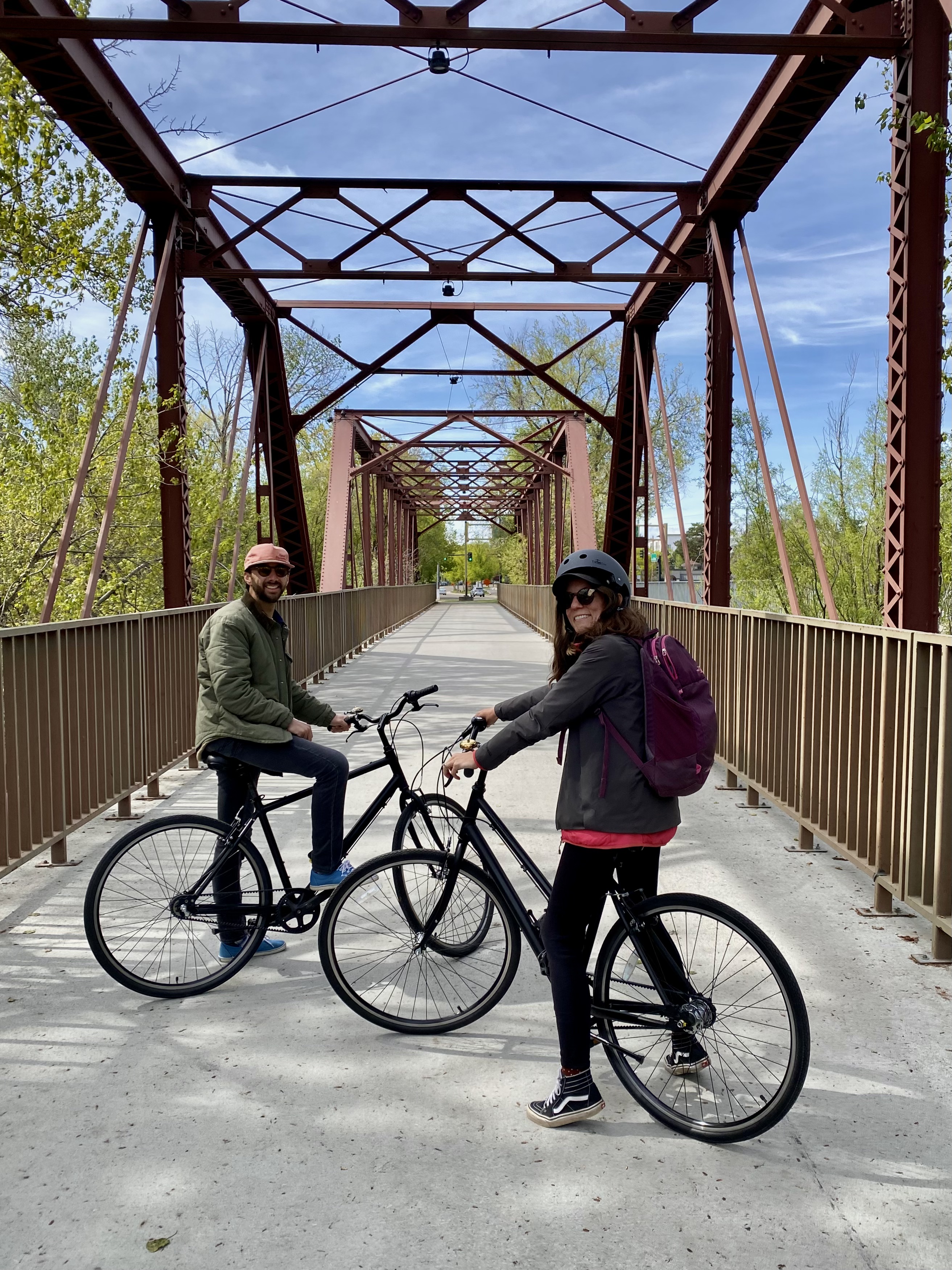 Caroline, in black pants and jacket, and jonathan, in a green jacket and pink hat, sit on black bicycles. behind them is a metal bridge. 