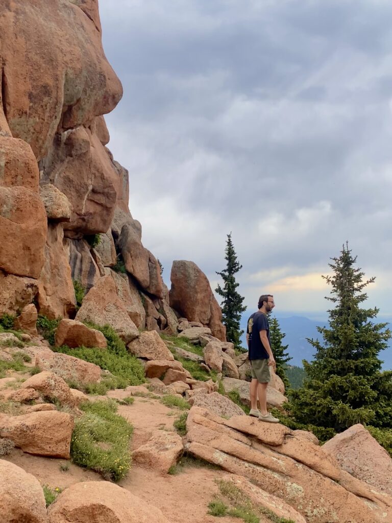 Jonathan, in a black shirt and tan shorts, stands on a red rock by a green pine tree.