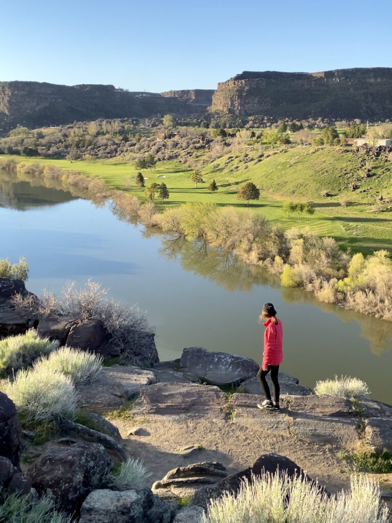 Caroline, in a pink jacket and black pants, looks out across a green river with bright green valley and blue sky background. 