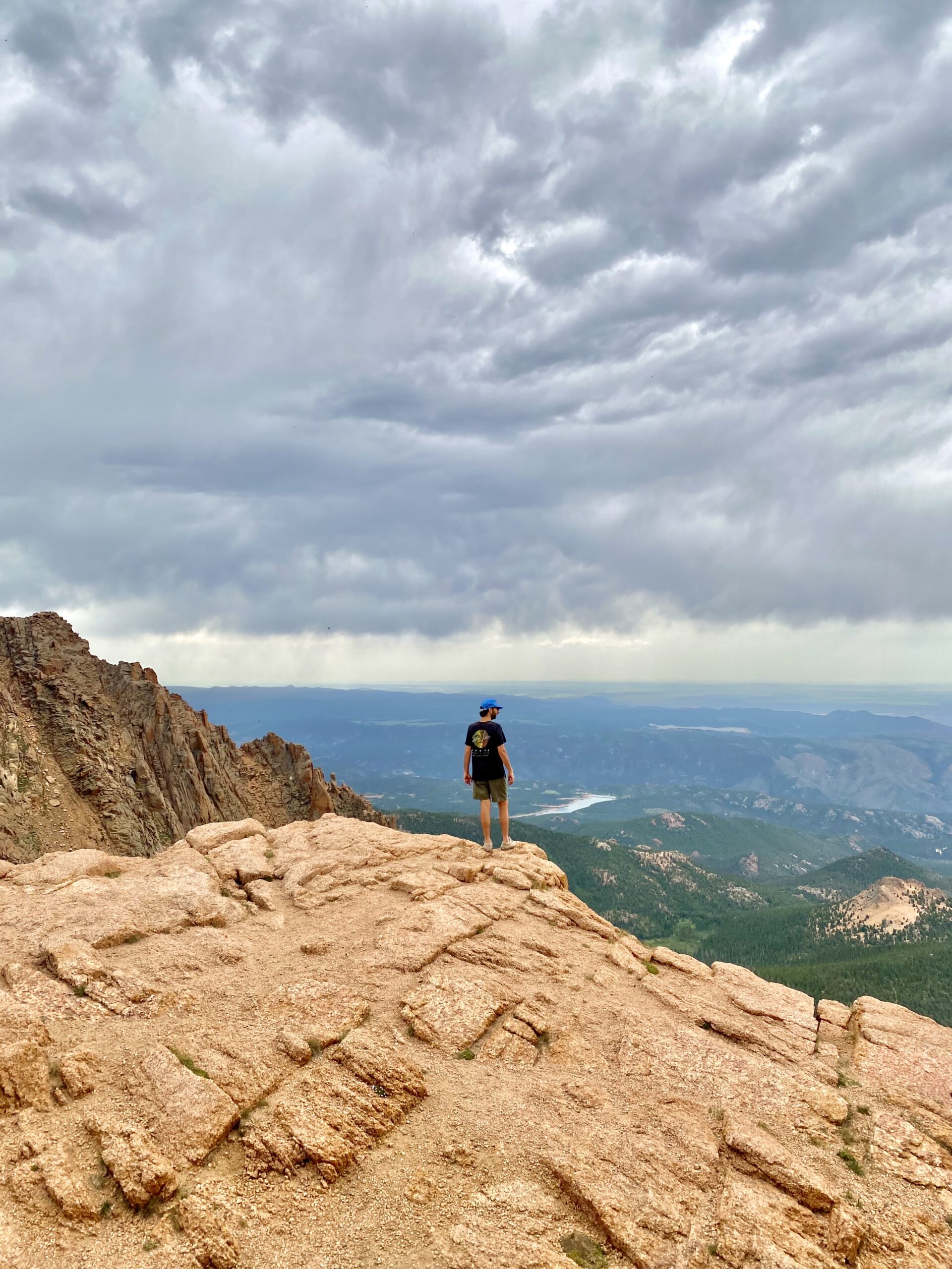 Jonathan, in a black shirt and blue hat, stands on the edge of a red rock. Dark & stormy clouds form behind him. 