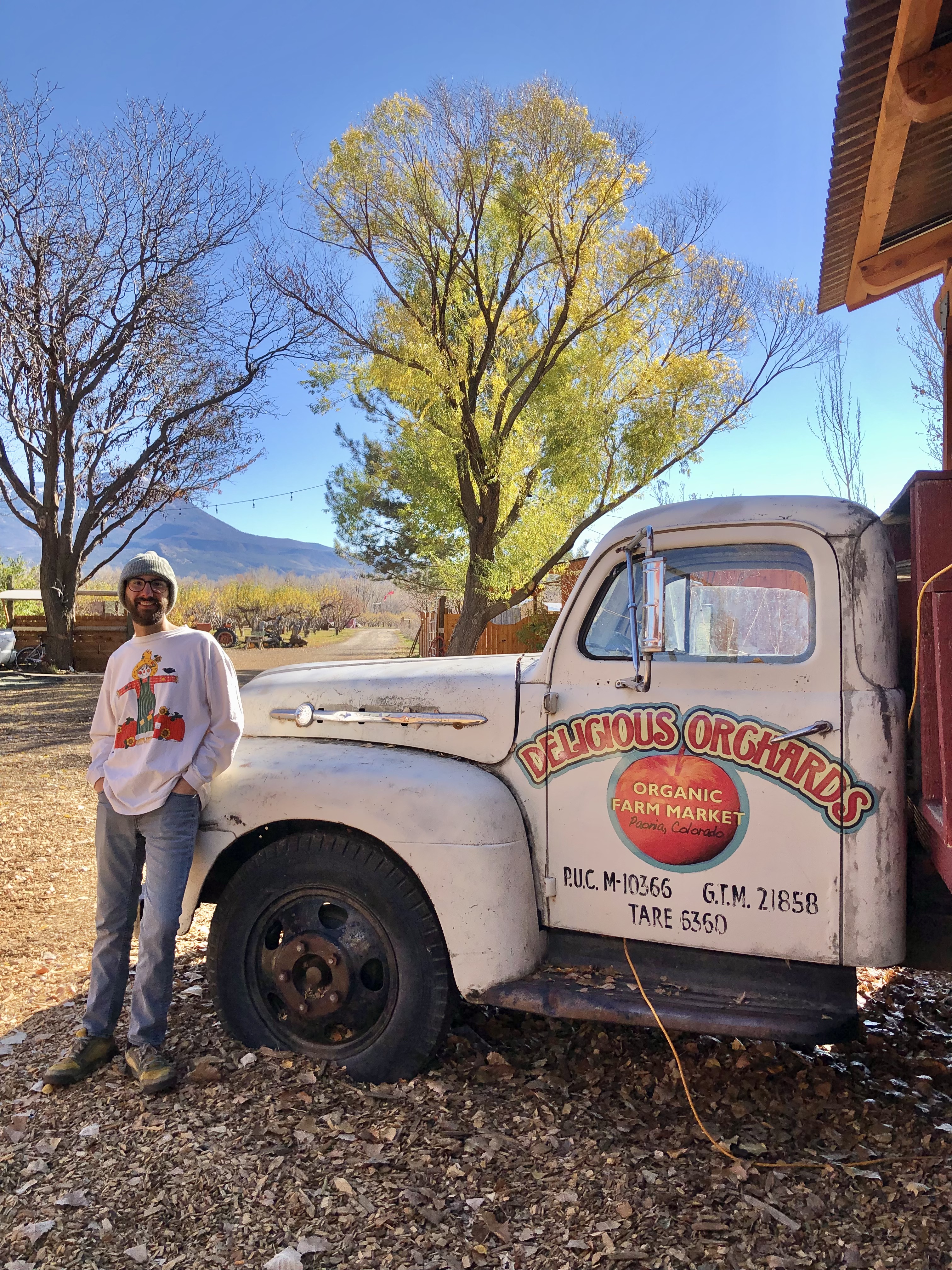 Jonathan, in a white shirt and green beanie, stands next to a white truck that states "delicious orchards". small towns to explore on a colorado road trip.